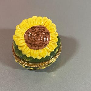 Puede incluir: Un joyero redondo de cerámica con una tapa en forma de girasol esculpido, con pétalos de color amarillo brillante y un centro marrón texturizado. La base está pintada de verde con detalles de hojas y rodeada por una bisagra y cierre de metal dorado.