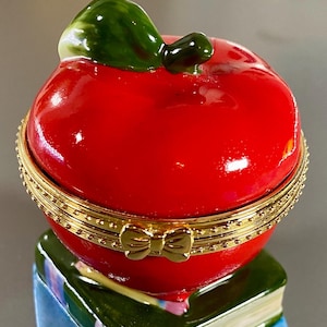 Puede incluir: Una caja de joyas de manzana roja con una hoja verde y un borde dorado. La caja está cerrada y tiene un pequeño lazo dorado en la parte delantera.