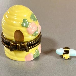 May include: A yellow ceramic beehive trinket box with a floral design and a small bee figurine. The box has a hinged lid and a brass clasp.
