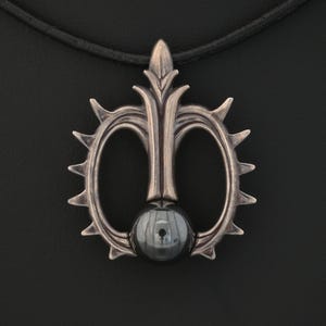 May include: A silver-toned pendant necklace with a unique design. The pendant features a central dark gray sphere, surrounded by an ornate, symmetrical shape with pointed edges and a decorative top. The necklace has a black cord.