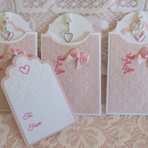 May include: Three pink gift tags with a white background and a heart pattern. Each tag has a silver charm with a heart design hanging from the top. The tags have the text "Just for You" written in pink. One tag has a blank white space with the words "To:" and "From:" written in pink.