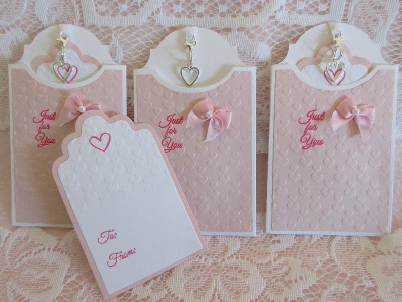May include: Three pink gift tags with a white background and a heart pattern. Each tag has a silver charm with a heart design hanging from the top. The tags have the text "Just for You" written in pink. One tag has a blank white space with the words "To:" and "From:" written in pink.