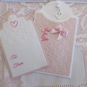 May include: A pink and white gift tag with a heart design and the text "Just for You". The tag has a small silver charm in the shape of a heart.