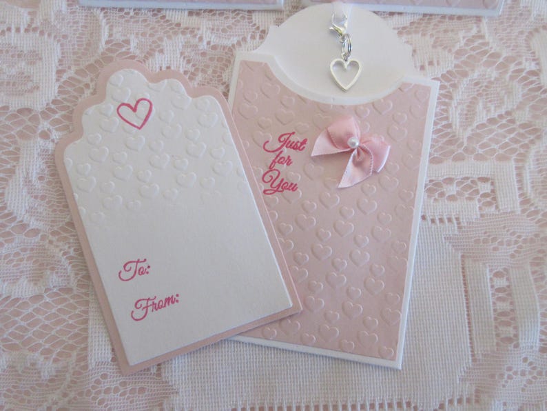 May include: A pink and white gift tag with a heart design and the text "Just for You". The tag has a small silver charm in the shape of a heart.