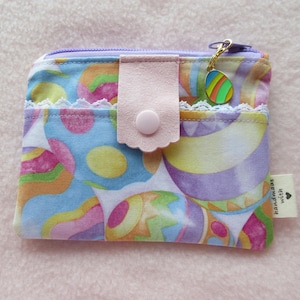 May include: A small purple and white fabric pouch with a pink flap and a zipper closure. The pouch has a colorful egg pattern and a small charm with a colorful egg design. The pouch has a label that says "handmade with love".