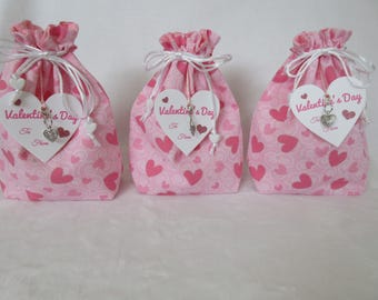 May include: Three pink fabric gift bags with a heart pattern. Each bag has a white heart-shaped tag that reads "Valentine's Day" and a small charm. The bags are tied with white ribbon.
