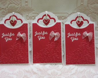 May include: Three red cardstock tags with a white scalloped border and a white heart charm. Each tag has the text "Just for You" in white script font and a white satin bow.