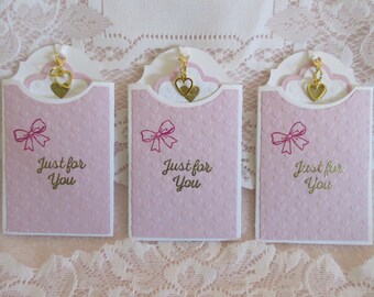 May include: Three pink cards with a gold heart charm hanging from the top. Each card has a pink bow and the text "Just for You" in gold lettering.