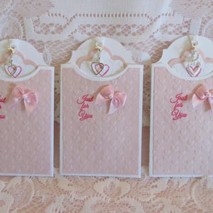 May include: Three pink and white cards with a heart-shaped opening at the top. Each card has a silver charm hanging from the opening, and a pink bow on the front. The cards have the text "Just for You" written on them.