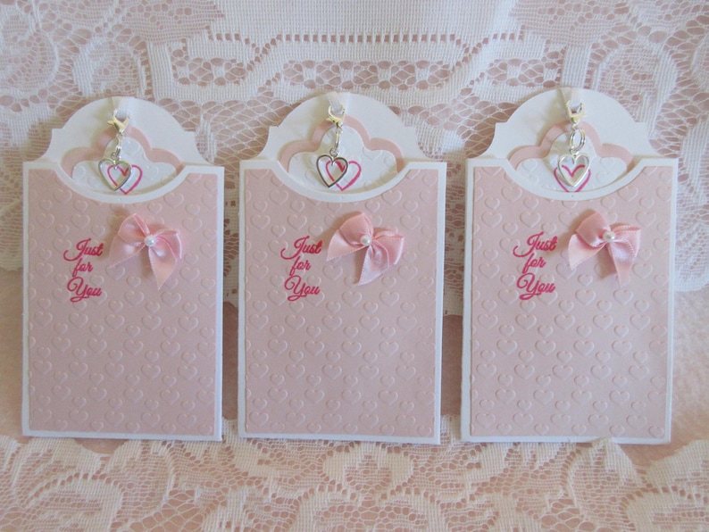May include: Three pink and white cards with a heart-shaped opening at the top. Each card has a silver charm hanging from the opening, and a pink bow on the front. The cards have the text "Just for You" written on them.