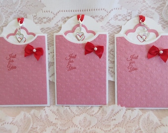 May include: Three pink cardstock gift tags with a white border and a heart pattern. Each tag has a silver charm with two hearts, a red bow, and the text "Just for You".
