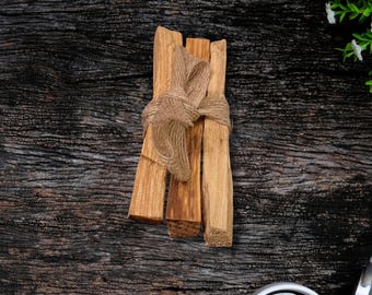 Puede incluir: Un paquete de cuatro palitos de Palo Santo marrones atados con cuerda sobre un fondo de madera oscura.