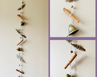 May include: A white string with cinnamon sticks, feathers, and small gold bells hanging from it. The string is hanging from a wall.