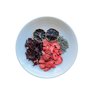 Puede incluir: Un cuenco de cerámica blanco lleno de ingredientes secos. El cuenco contiene fresas secas, hibisco, lavanda y dos mitades de cítricos secos. Los ingredientes están dispuestos en secciones.