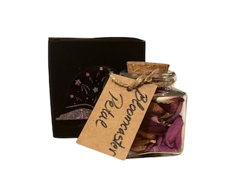 May include: A small glass jar filled with dried pink and purple flower petals, labeled "Bloomcaster Petal." The jar has a cork stopper and is tied with twine to a brown paper tag. A black box with a glittery design is in the background.