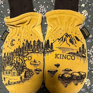 Puede incluir: Un par de mitones de cuero amarillo con puños negros. Cada mitón presenta una ilustración en blanco y negro de una cabaña junto al lago y montañas con la palabra "KINCO". Los mitones están etiquetados como "Heatkeep".