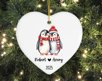 May include: White heart-shaped Christmas ornament featuring two penguins wearing scarves and hats. The ornament has the names "Robert" and "Avery" with a heart symbol, and the year "2025" printed on it. The ornament hangs from a gold string.