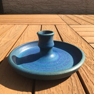 Peut inclure: Un bougeoir en céramique fait à la main dans les tons de bleu. Le bougeoir comporte une coupelle centrale pour la bougie et une base large et peu profonde. La pièce est glacée avec une finition mouchetée et texturée, et repose sur une surface en bois.
