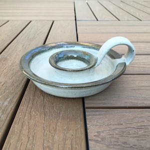 Peut inclure: Un bougeoir en céramique fait à la main, avec une base circulaire et un puits central pour la bougie. Le bougeoir a une anse incurvée et est fini avec un glaçage bleu clair et une bordure marron. Sur une surface en bois.
