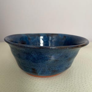 May include: A handmade ceramic bowl with a deep blue glaze. The bowl has a rounded shape with a slightly flared rim and a reddish-brown base. The blue glaze has variations in colour, giving it a textured appearance.