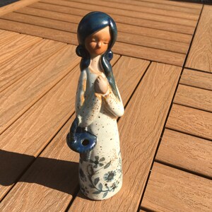 May include: A ceramic figurine of a woman with a blue head and hair, wearing a white dress with blue floral patterns. The figurine has a blue purse and is standing on a wooden surface.