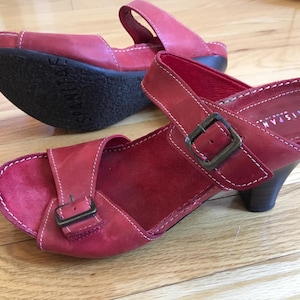 May include: Red leather sandals with a small heel. The sandals feature a toe strap and an ankle strap with a buckle. White stitching details the edges. The sole is black. The brand name 'Hispanias' is visible.