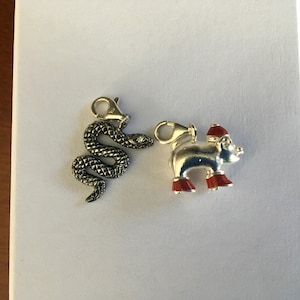 May include: Two metal charms: a detailed snake with a small gem and a pig wearing a red hat and boots. The charms are silver-toned with black detailing on the snake and red enamel on the pig.
