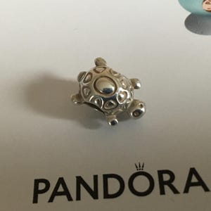 May include: A silver turtle charm with a detailed shell and heart-shaped cutouts. The charm is a small, decorative accessory, likely for a bracelet or necklace. The charm is on a white surface with the word PANDORA visible.