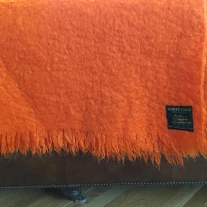 May include: A vibrant orange fuzzy throw blanket with a textured, shaggy appearance and a fringed bottom edge. A black rectangular label is stitched onto the lower right corner, featuring the white text Robert Burns and additional small print.