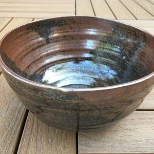 May include: A handcrafted ceramic bowl with a rustic, earthy appearance. The bowl has a rich brown hue with dark, textured details and a glossy interior. The bowl is suitable for serving salads or displaying fruit.