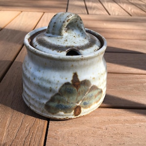 May include: A ceramic sugar bowl with a lid and handle. The bowl is off-white with brown and green floral designs. The lid has a small handle and a hole for a spoon. The bowl is sitting on a wooden surface.