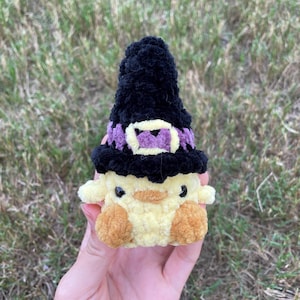 Puede incluir: Un pollito de crochet amarillo con un sombrero de bruja negro con una banda morada. El pollito tiene ojos negros y un pequeño pico naranja.