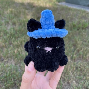 May include: A black crocheted plush toy with a blue crocheted hat. The toy has a pink nose and black eyes.