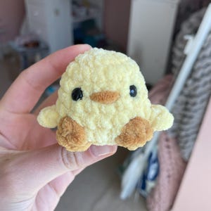 May include: A small yellow crocheted chick with brown feet and black eyes. The chick is being held in a hand.