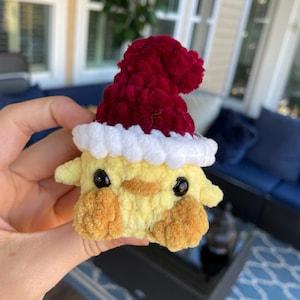 May include: A handmade, yellow crocheted chick wearing a red Santa hat with a white trim. The chick has black eyes and brown accents on its cheeks and beak. This festive plush toy is perfect for holiday decor or as a unique gift.