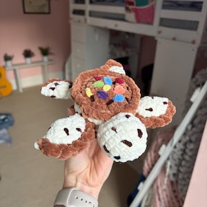 May include: A plush crochet turtle toy featuring a brown shell with colorful, multi-colored embroidered accents. The turtle has a white head and flippers, each decorated with small, dark brown spots, crafted from a soft, textured yarn material.