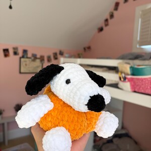 May include: A handmade crochet plush toy shaped like a dog with a white head, black ears, and a black nose. The body is made of bright orange yarn, and the four paws are white. The plush has a soft, textured appearance with small black eyes.