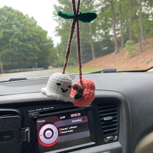 May include: A handmade car charm featuring a white crocheted ghost with a smiling face and a crocheted red apple with a brown stem. The charm hangs from a brown string with a green leaf accent.