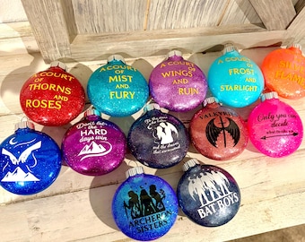May include: A selection of glittery Christmas baubles in various colours, each with text. The baubles feature phrases like "A Court of Thorns and Roses" and "Bat Boys". The baubles are round with silver tops.