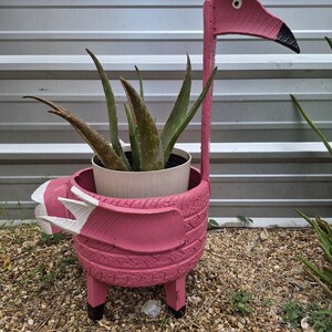 Pode incluir: Um vaso em forma de flamingo cor-de-rosa feito de um pneu reciclado, com um pescoço comprido, um bico com ponta preta e detalhes de asas brancas. O vaso contém uma planta de aloé vera com folhas verdes pontiagudas e assenta em quatro pernas.