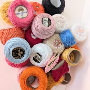May include: A selection of colourful thread spools in shades of pink, orange, blue, white, and yellow. Some spools have labels with text like "Pearl Cotton" and "DMC". Suitable for crafting and embroidery.