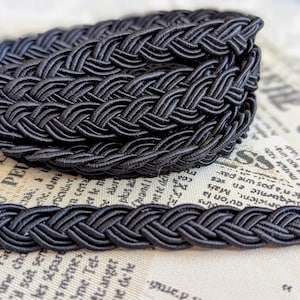 Puede incluir: Primer plano de un ribete trenzado negro, tejido apretadamente y enrollado. El ribete es de un negro brillante y se encuentra sobre un fondo de periódico vintage. Adecuado para proyectos de artesanía y costura.