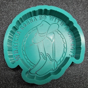 May include: A teal silicone mold featuring a circular design with a cowboy hat resting on a pair of cowboy boots. A lasso encircles the boots and hat. The text "WHATCHA GONNA DO WITH A COWBOY?" is embossed in a circular pattern around the central image.