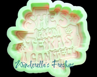 May include: A light green silicone mold featuring the text "SHE'S FROM TEXAS I CAN TELL" in a retro font, accompanied by small flower icons. The mold is shaped like the state of Texas and includes the text "Winderella's Freshies" at the bottom.