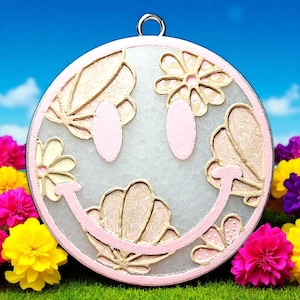 May include: A round, pink and white smiley face pendant with floral and shell designs. The pendant has a silver loop at the top for hanging. The background is a bright blue sky and green grass with colorful flowers.