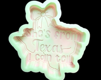 May include: A pale green cookie cutter shaped like the state of Texas with a bow at the top. The interior features the embossed text "She's from Texas I can tell." The cutter has a raised edge and a smooth, light-colored plastic finish.