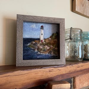 May include: A framed artwork depicts a lighthouse on a rocky shore, with a blue sky and ocean. The lighthouse is white with a red top. The frame is a light gray color. The artwork is displayed on a wooden shelf.