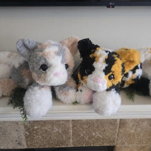 Peut inclure: Deux jouets en peluche en forme de chat avec une fourrure douce et texturée. Le chat de gauche présente un mélange de taches grises, blanches et fauves. Le chat de droite affiche un motif calicot noir, orange et blanc. Yeux sombres et museaux roses.