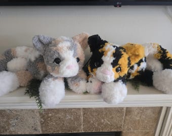 May include: Two plush cat toys with soft, textured fur. The left cat features a mix of grey, white, and tan patches. The right cat displays a calico pattern of black, orange, and white. Both have large dark eyes, pink noses, and white paws.