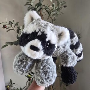 Puede incluir: Un peluche de mapache con pelaje suave y texturizado en tonos blanco, gris y negro. El animal de peluche tiene una máscara negra distintiva alrededor de los ojos, una nariz negra y una cola rayada. El pelaje se ve esponjoso y multicolor.
